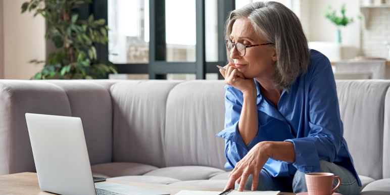 A grey-haired middle-aged woman in a sky-blue shirt sitting on a couch and looking into her laptop.