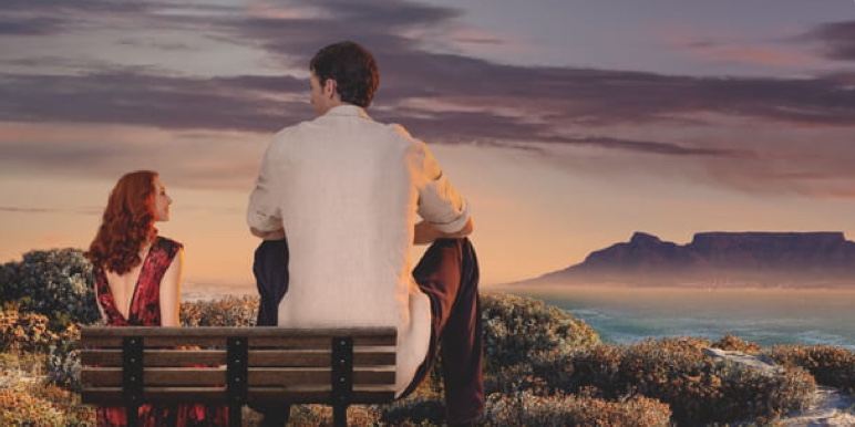 #BornWithMomentum. A very tall man sitting side by side with a smaller woman on a bench overlooking the sea, representing support given to clients to achieve their financial goals at every step of their life journey.