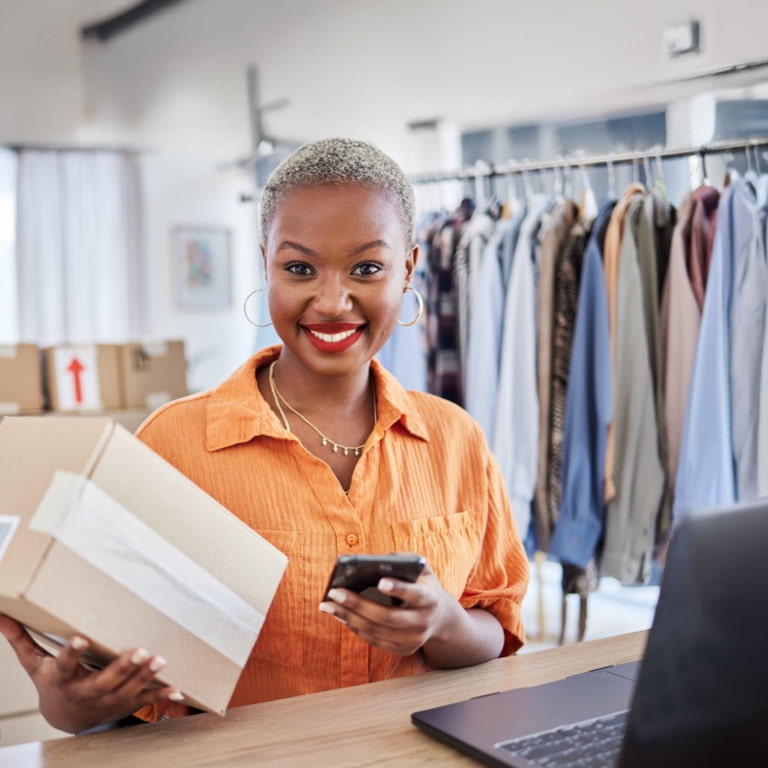 Smiling female business owner in an orange shirt holding a package and using a smartphone in a boutique clothing store. Shelves with boxes and racks of clothes suggest a thriving retail business, secured with business insurance.