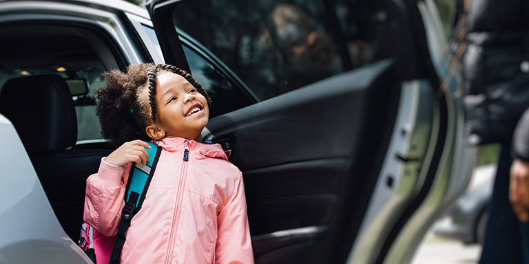 A happy little girl in a pink rain jacket, carrying a school bag, standing outside of an open car door, confident that she is safe and secure.