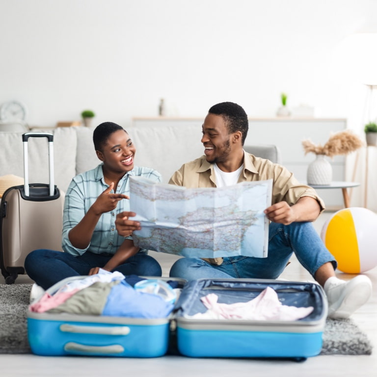 A young African couple looking at a paper map, smiling confidently that they have all their items packed and itinerary planned for their December holiday.