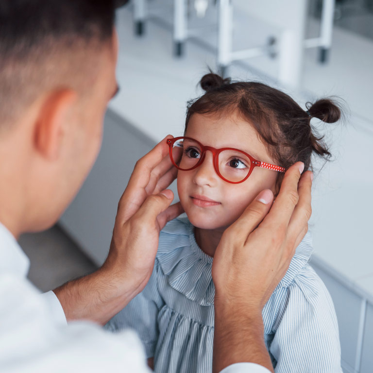 A cute young girl smiles while an optometrist holds out red-framed glasses for her to try on.