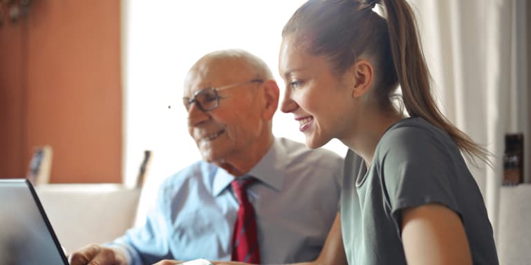A lady and an elderly man sitting in front of a laptop and smiling with bright daylight coming from the window next to them.