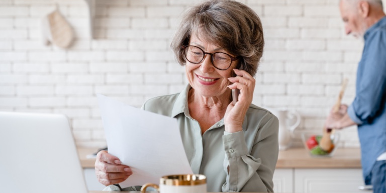 A smiling retired, elderly woman on her mobile phone, reviewing her policy details online and holding a hardcopy of her contract now managed by another insurer.