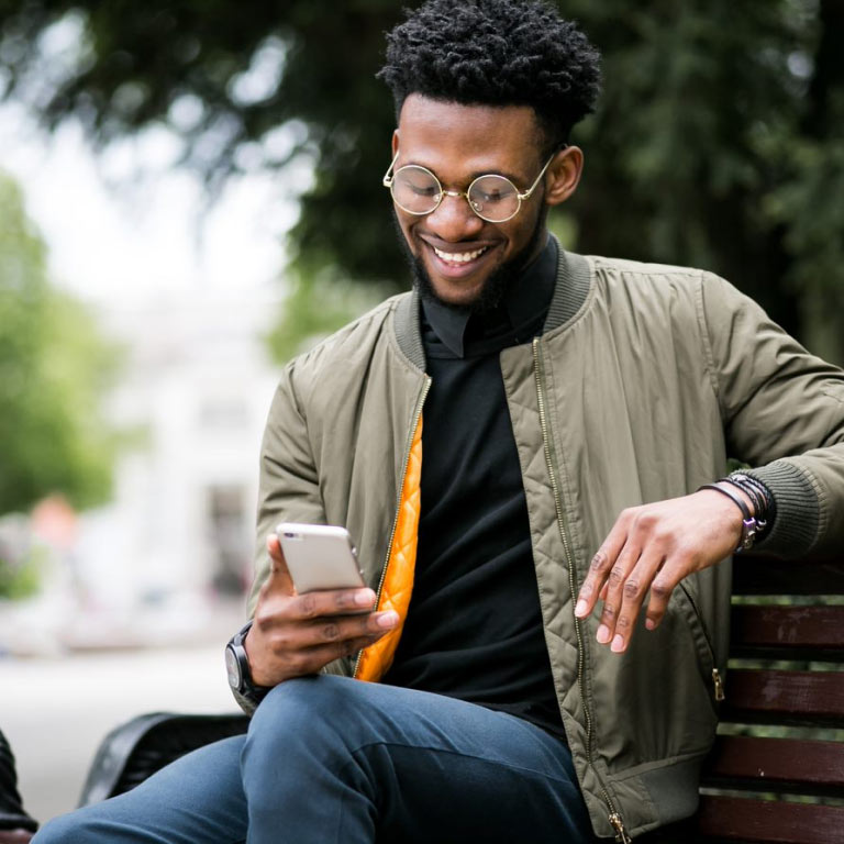 young man sitting on a bench outdoors, smiling while looking at his cell phone, reading up on Momentum’s Evolve medical aid option.