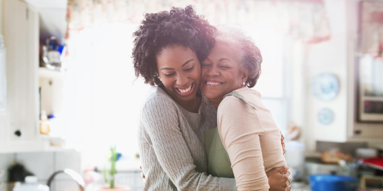 A mom hugs her young smiling daughter with eyes closed. They both are wearing yellow tops and have dark long hair.