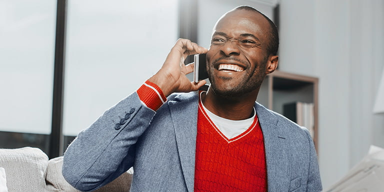 A man smiling broadly and speaking on his cell phone, wearing a grey blazer and a red top underneath.