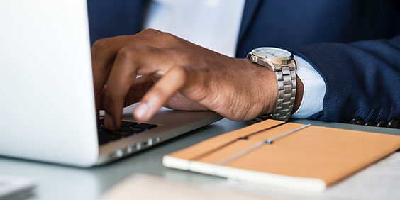 A faceless, close-up view of a financial adviser typing on his laptop with a notebook at his desk.