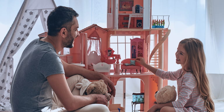 A little girl and her father are playing with her dollhouse on the floor. Alongside the house is a little tent.