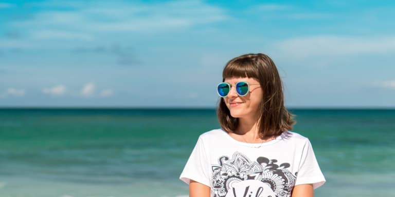 A brunette lady standing at a sunny beach wearing sunglasses and a black white t-shirt.