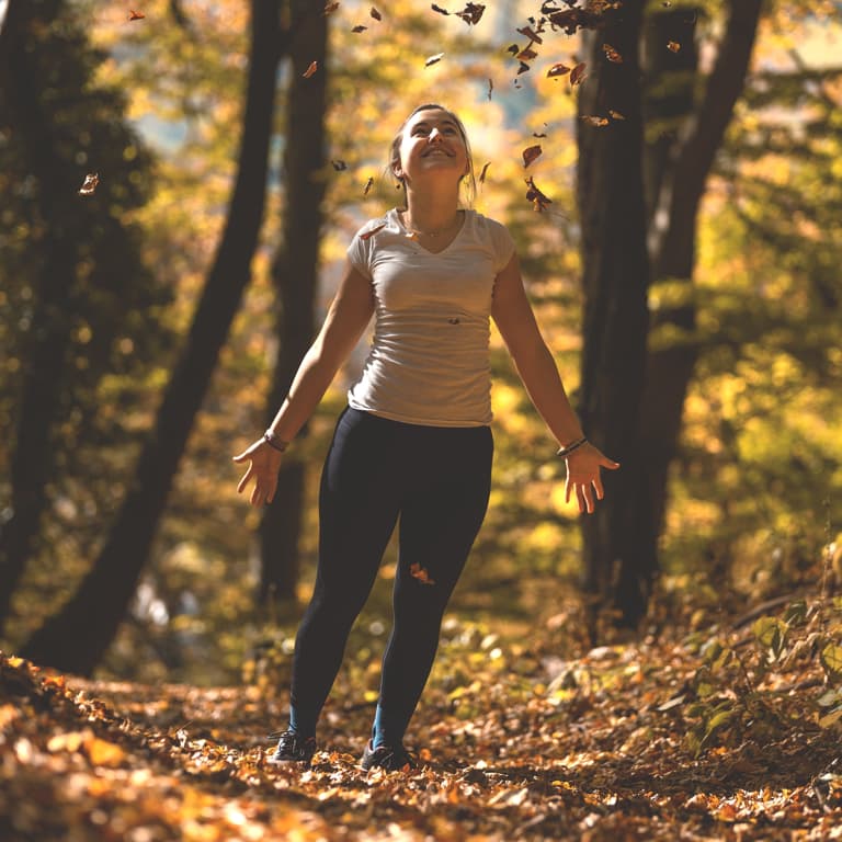 Relaxed woman walking through the woods looking up and smiling at the sun with arms open.