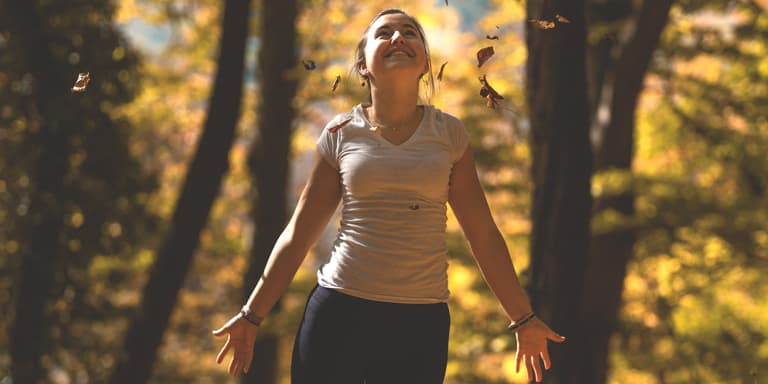 Relaxed woman walking through the woods looking up and smiling at the sun with arms open.