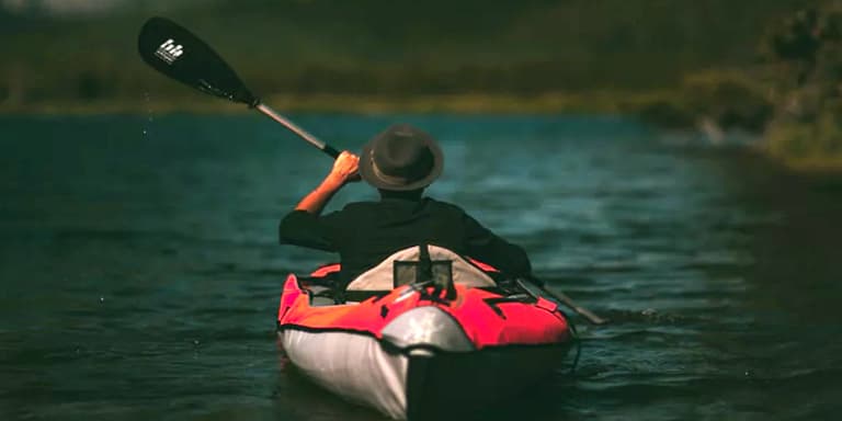 A man rowing a kayak in a dam, wearing a black shirt and a hat.