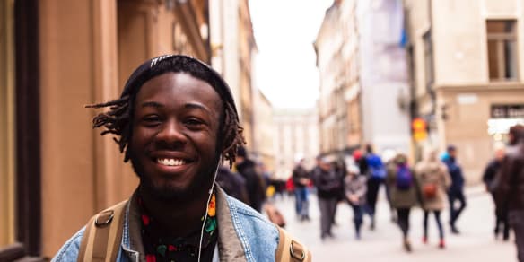 A young man with dreadlocks walking and smiling in a busy street while wearing a black beanie hat and earphones.