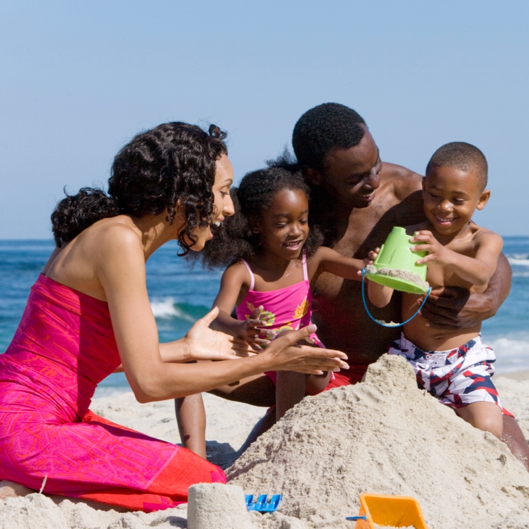 Mother and father happily playing on the beach with their two children in the sand building a sandcastle, near the ocean.