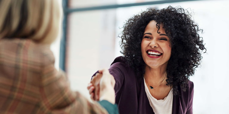 Two women in a modern style office smiling and shaking hands at a business meeting.