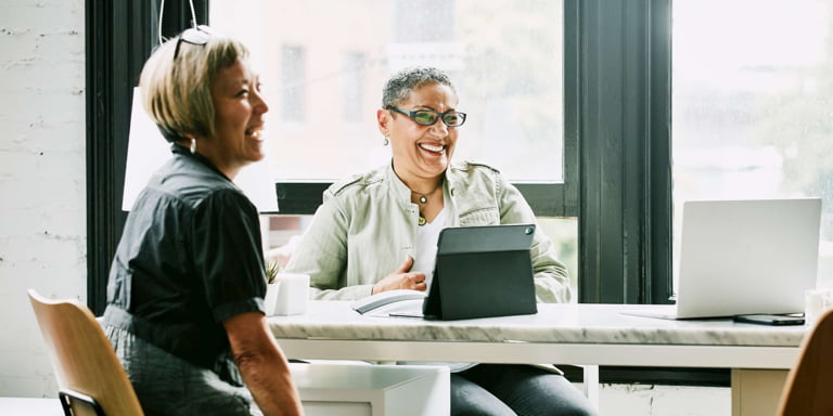 Two women sitting at a small round office table with their laptops are looking at, and laughing with another colleague.