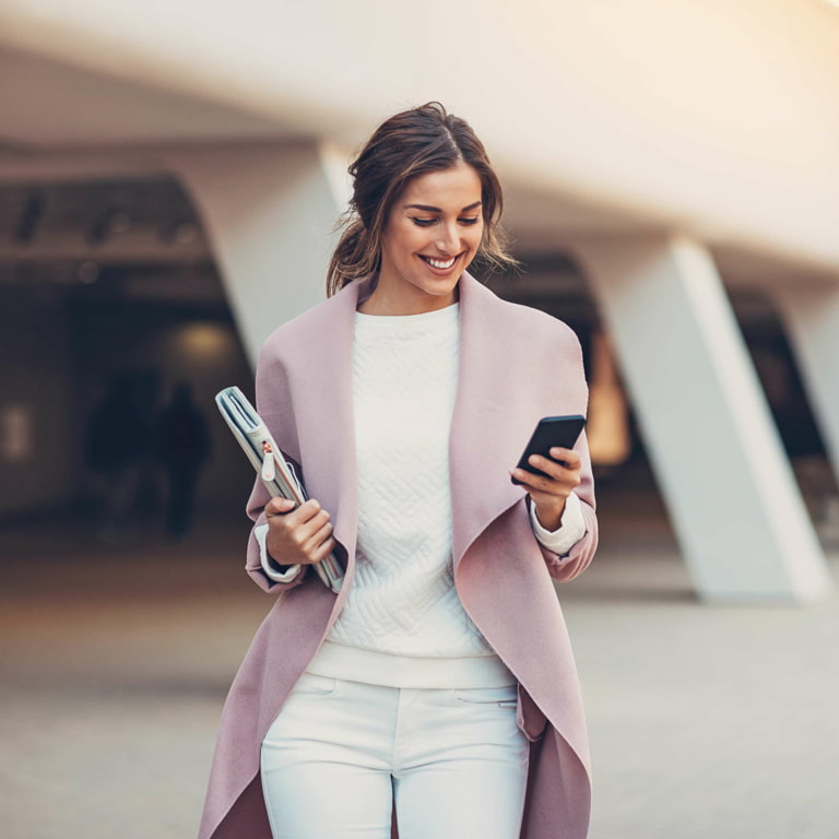 Lady wearing white jeans, white jumper, and a pink jacket walks out of a building holding her folder in one hand while looking at her cell phone reading up about Business Continuity Assurance products for her business.
