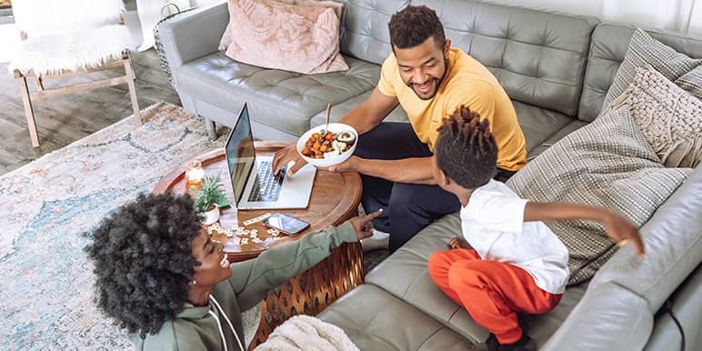 A small, happy family of three interacting in a living room as dad eats his lunch and mom plays with the kid.