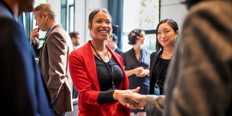 A woman in a red suit and black shirt, shaking hands with a man in a brown suit at a conference.