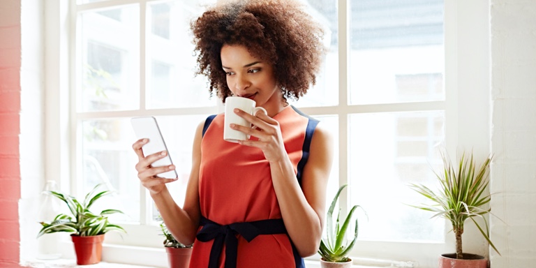 A woman in a red shirt, with a coffee mug in her hand, standing next to a glass wall while typing on her cell phone.