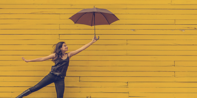 Young girl holding a red umbrella against a yellow wall. She’s smiling and is confident about her journey to success.