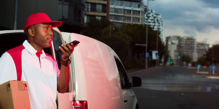 A courier stands in the city, speaking on his phone while holding a box, ready to make a delivery.