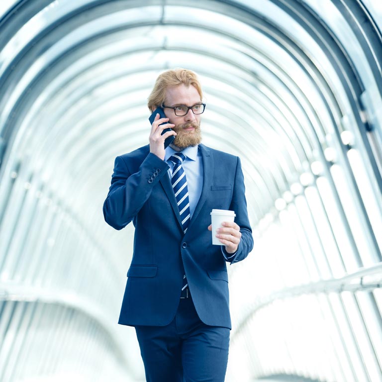 A man with ginger hair and beard in a dark blue suit is walking while holding a cup of coffee and holding a phone to his ear.