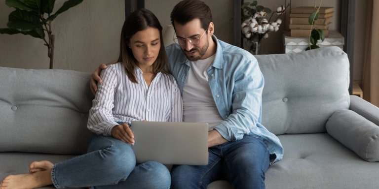 A couple in jeans seating on a grey couch and working on a laptop together.