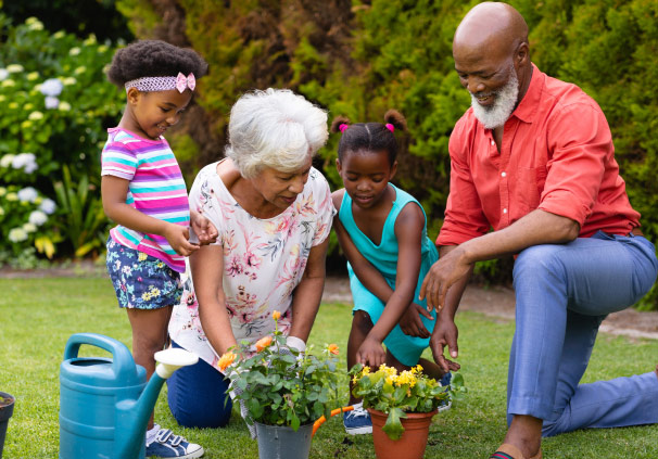 A Retired couple in their garden, showing their two granddaughters how to water two pots of flowers.