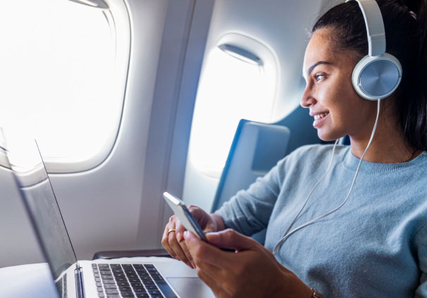 A happy young woman on an airplane with electronics and headphones looking outside of the window.