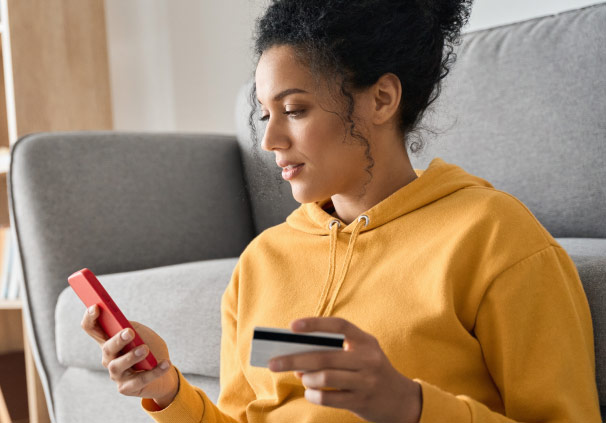 A young woman at home, sitting on the floor, accessing her digital savings account on her mobile phone with her Momentum Money card in hand. 