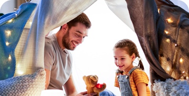 Father and his young daughter sitting under a makeshift tent in their living room. They're smiling while the father holds a small teddy bear while telling her a story.