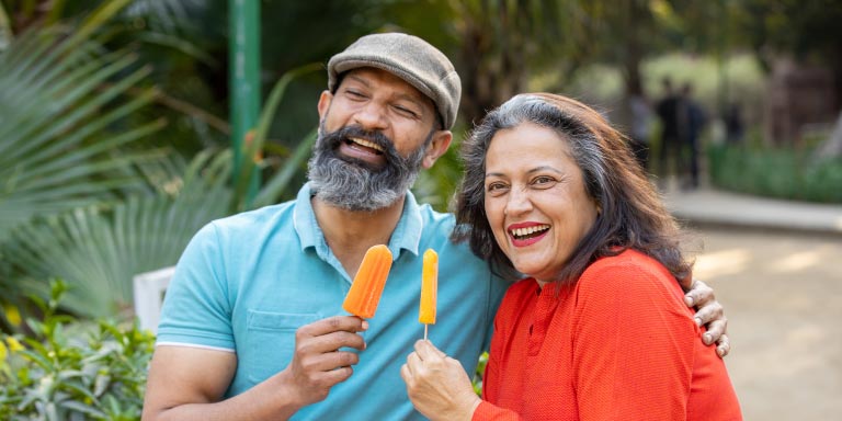 Happy Indian senior couple eating ice lollies in a park outdoor, old mature couple enjoying retirement life.